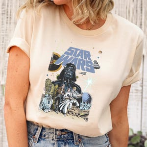 May include: Cream-colored Star Wars graphic tee featuring Darth Vader and other characters. The shirt has a classic crew neck and short sleeves. The graphic is in shades of blue, black, and beige. The model is wearing light blue ripped jeans.