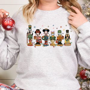 May include: A light gray sweatshirt featuring a colorful graphic of Disney characters dressed as nutcrackers. The characters include Mickey Mouse, Minnie Mouse, Donald Duck, and Goofy. A red Christmas ornament is held in the hand.