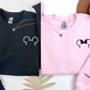 以下が含まれることがあります： 白のミッキーマウスとミニーマウスの耳のデザインが施された黒とピンクのスウェットシャツ。各スウェットシャツには金のネックレスと、袖にはローマ数字のXLXVIIIMMXXが付いています。左上には「Personalized Gift EMBROIDERED SWEATSHIRT & HOODIE」と書かれた丸いロゴがあります。