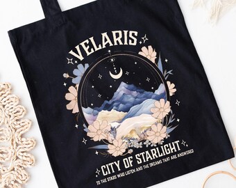May include: Black tote bag with a graphic design featuring the text "VELARIS CITY OF STARLIGHT" and a mountain landscape with flowers and a crescent moon. The bag has black handles.