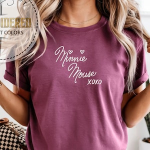 May include: A maroon t-shirt with the words "Minnie Mouse" in white script, with two small hearts and "XOXO" underneath. The shirt has rolled-up sleeves and is part of a personalized gift collection.