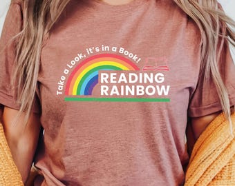 May include: A mauve-colored t-shirt with the text "Take a Look, it's in a Book! READING RAINBOW" in a rainbow design. The shirt is made of soft material and is suitable for casual wear. The shirt is paired with blue jeans.