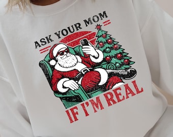 May include: White sweatshirt with a graphic of Santa Claus taking a selfie with a Christmas tree. The text reads "ASK YOUR MOM IF I'M REAL" in red and black.
