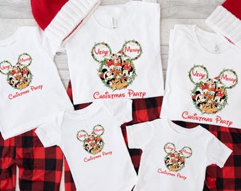 May include: A collection of white t-shirts and a onesie with a Mickey Mouse-themed design, paired with red and black plaid pajama pants and red and white knit hats. The shirts and onesie feature a wreath design with the words "Very Merry Christmas Party".