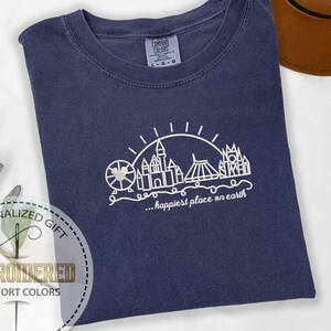 May include: Navy blue Comfort Colors t-shirt with a white line drawing of a theme park skyline, including a Ferris wheel, castle, and other structures. The text "...happiest place on earth" is below the drawing. The shirt is folded on a white surface.