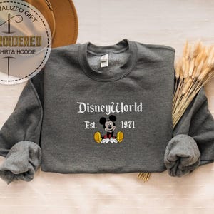 May include: A charcoal grey sweatshirt featuring the words "DisneyWorld Est. 1971" and a Mickey Mouse design. The top has long sleeves and a round neck. The image also includes a hat and dried flowers.