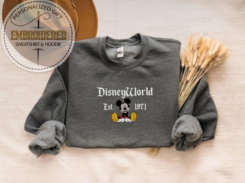 May include: A charcoal grey sweatshirt featuring the words "DisneyWorld Est. 1971" and a Mickey Mouse design. The top has long sleeves and a round neck. The image also includes a hat and dried flowers.