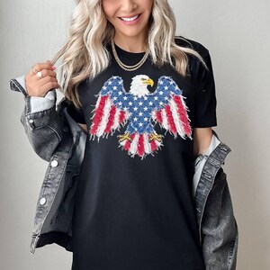 May include: A black short-sleeved t-shirt featuring a graphic of a bald eagle with wings spread. The eagle design incorporates a red, white, and blue American flag pattern with a distressed, frayed texture. The shirt is worn under a gray denim jacket.
