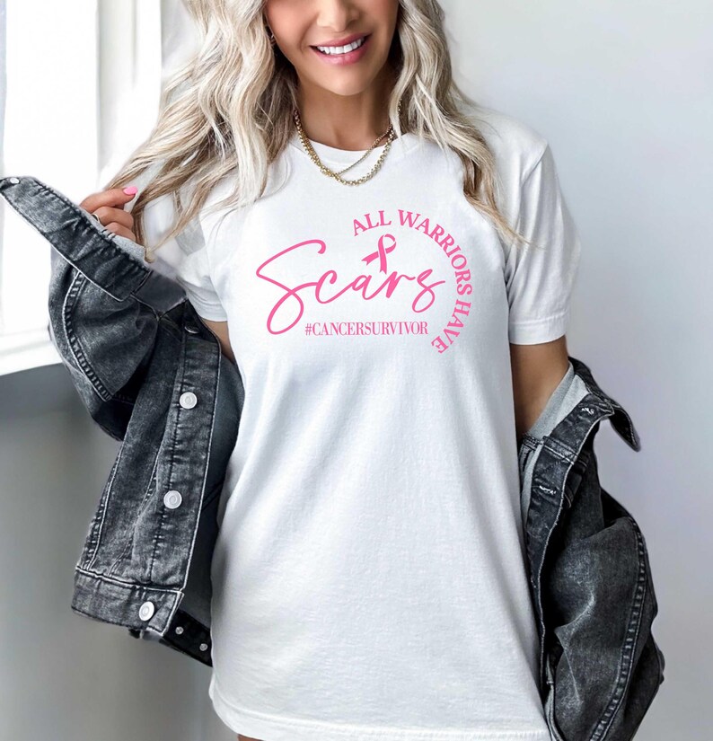 May include: White t-shirt with a pink ribbon and the text "All Warriors Have Scars #cancersurvivor" printed on the front.