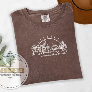 May include: A brown Comfort Colors t-shirt with a white graphic of a Ferris wheel, castle, and other structures, with the text "...happiest place on earth". The shirt is folded on a white surface.