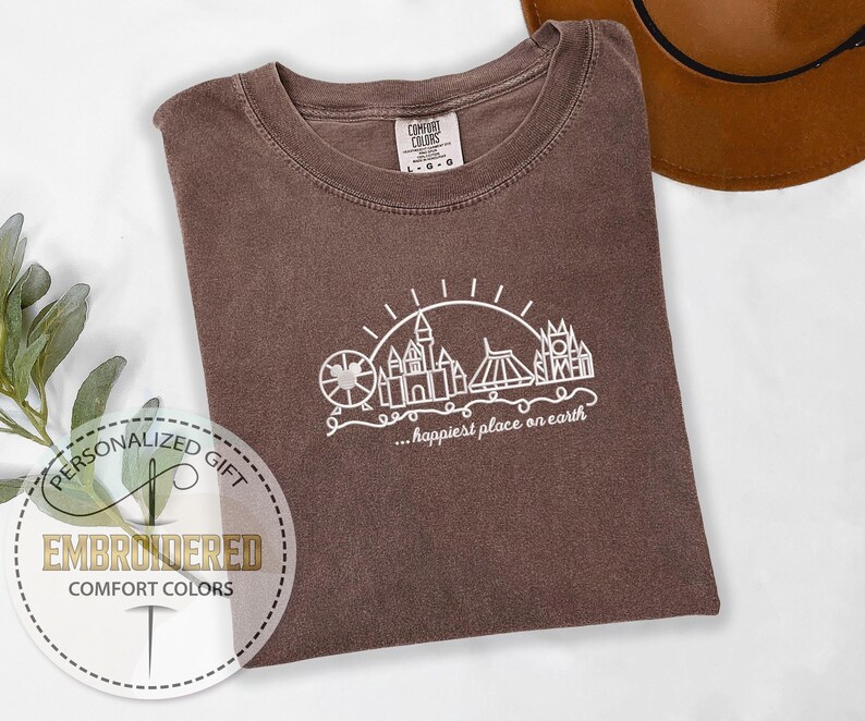May include: A brown Comfort Colors t-shirt with a white graphic of a Ferris wheel, castle, and other structures, with the text "...happiest place on earth". The shirt is folded on a white surface.