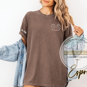 May include: A brown t-shirt featuring an embroidered Sally face design on the chest. The left sleeve is embroidered with the words "His Sally". The shirt is part of the "Comfort Colors" collection.