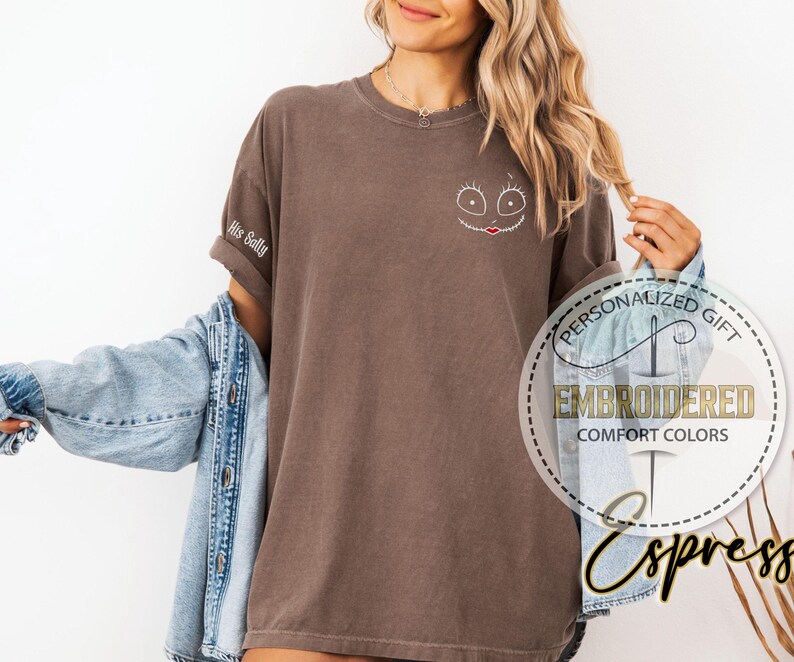 May include: A brown t-shirt featuring an embroidered Sally face design on the chest. The left sleeve is embroidered with the words "His Sally". The shirt is part of the "Comfort Colors" collection.