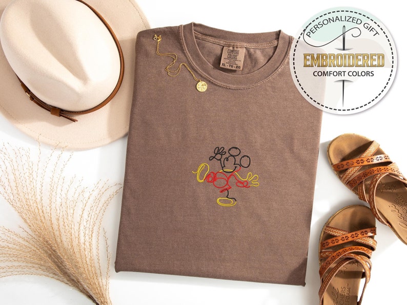 Op de afbeelding: Een bruin T-shirt met een eenvoudig geborduurd ontwerp van Mickey Mouse in zwart, rood en geel garen. Het T-shirt is gevouwen en plat gelegd op een wit oppervlak. De tekst "Gepersonaliseerd cadeau" en "Geborduurd Comfort Colors" is zichtbaar in de rechterbovenhoek van de afbeelding.