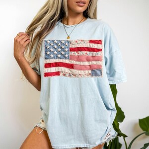 May include: Light blue t-shirt featuring a patchwork American flag design. The flag incorporates various fabrics, including denim, floral, and gingham patterns, with white stars on a blue background and red and white stripes.