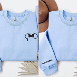 以下が含まれることがあります： 黒いミッキーマウスの耳のデザインが施された、2つの水色のスウェットシャツ。1つはリボン付き、もう1つはリボンなし。各スウェットシャツの袖には、黒で「XLVII.MMXX」のローマ数字がプリントされています。白い表面に表示されています。