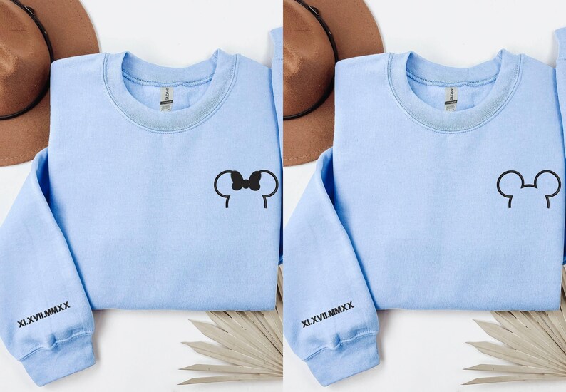 以下が含まれることがあります： 黒いミッキーマウスの耳のデザインが施された、2つの水色のスウェットシャツ。1つはリボン付き、もう1つはリボンなし。各スウェットシャツの袖には、黒で「XLVII.MMXX」のローマ数字がプリントされています。白い表面に表示されています。