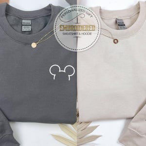 以下が含まれることがあります： チャコールグレーとベージュの2枚のスウェットシャツが表示されています。それぞれ前面に白いミッキーマウスの頭のデザインがあります。各スウェットシャツの左袖にはローマ数字があります。画像には「PERSONALIZED GIFT EMBROIDERED SWEATSHIRT & HOODIE」というテキストが含まれています。