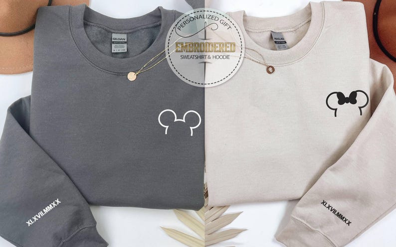 以下が含まれることがあります： チャコールグレーとベージュの2枚のスウェットシャツが表示されています。それぞれ前面に白いミッキーマウスの頭のデザインがあります。各スウェットシャツの左袖にはローマ数字があります。画像には「PERSONALIZED GIFT EMBROIDERED SWEATSHIRT & HOODIE」というテキストが含まれています。