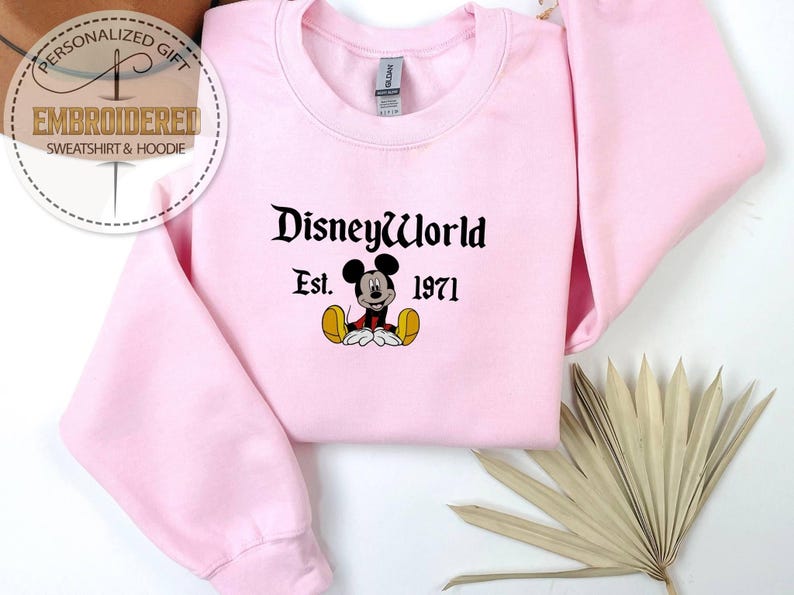 May include: A pale pink crew neck sweatshirt with the text "DisneyWorld Est. 1971" in black, and a Mickey Mouse graphic. The image also includes a decorative dried palm leaf. The sweatshirt is a casual style.