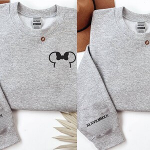 以下が含まれることがあります： 黒いミッキーマウスの耳のデザインが施された2つの灰色のスウェットシャツ。1つはリボン付き、もう1つはリボンなし。各スウェットシャツの袖にはローマ数字XLVII.MMXXがプリントされています。スウェットシャツは白い表面にあります。