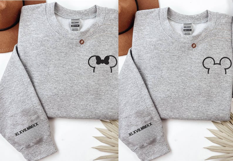 以下が含まれることがあります： 黒いミッキーマウスの耳のデザインが施された2つの灰色のスウェットシャツ。1つはリボン付き、もう1つはリボンなし。各スウェットシャツの袖にはローマ数字XLVII.MMXXがプリントされています。スウェットシャツは白い表面にあります。