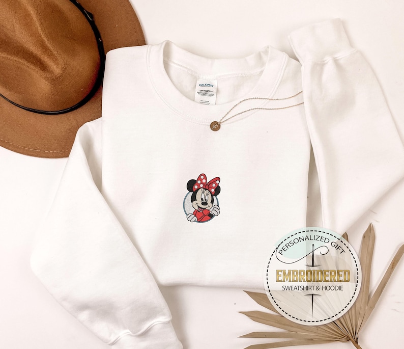 May include: White sweatshirt with a red and black embroidered image of Minnie Mouse wearing a red bow. The sweatshirt has a crew neck and long sleeves. The text "Personalized Gift" and "Embroidered Sweatshirt & Hoodie" is on a gold circle.