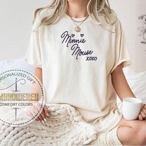 May include: Cream-colored t-shirt with the text "Minnie Mouse xoxo" in dark blue script. The shirt is part of the "Comfort Colors" collection and is embroidered. A person is holding a white mug.