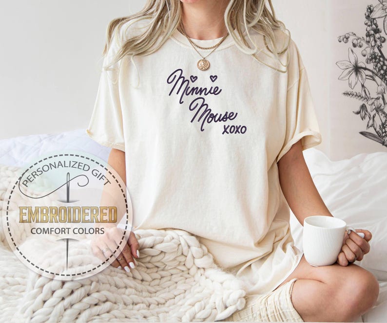 May include: Cream-colored t-shirt with the text "Minnie Mouse xoxo" in dark blue script. The shirt is part of the "Comfort Colors" collection and is embroidered. A person is holding a white mug.