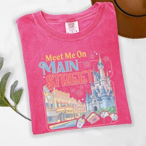 May include: A pink t-shirt with the text "Meet Me On Main Street" and a colorful graphic of a castle, buildings, and fireworks. The shirt is a Comfort Colors brand, size L-G.