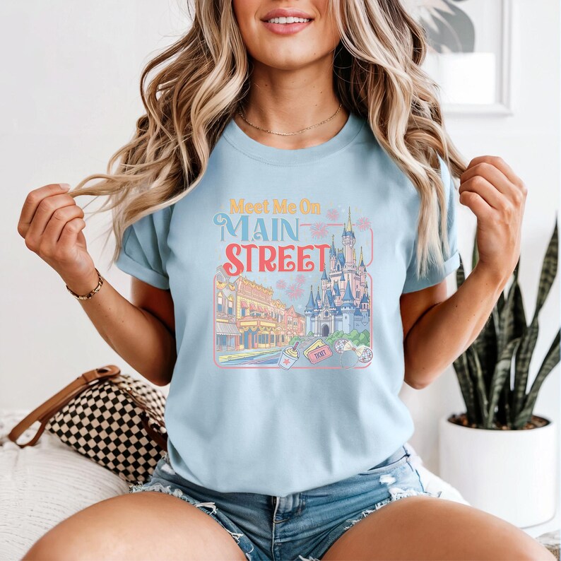 May include: Light blue t-shirt with a colorful graphic that says "Meet Me On Main Street." The graphic features a castle, buildings, and fireworks. The shirt is short-sleeved.