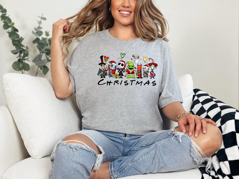 May include: A gray t-shirt with a graphic print of various cartoon characters from popular Christmas movies. The characters include Jack Skellington, Sally, Oogie Boogie, and the Grinch. The text "CHRISTMAS" is printed below the characters.