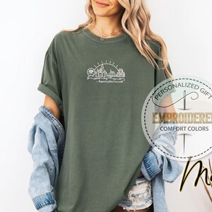 May include: Moss green t-shirt with a white embroidered amusement park design and the text "...happiest place on earth." The shirt is part of the "Comfort Colors" collection and is a personalized gift.