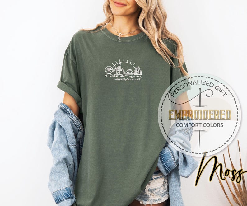 May include: Moss green t-shirt with a white embroidered amusement park design and the text "...happiest place on earth." The shirt is part of the "Comfort Colors" collection and is a personalized gift.