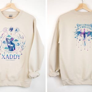 May include: A beige sweatshirt with a graphic print of a person with a dragon on the front and the text "XADDY" below. The back of the sweatshirt has a graphic print of a dragon with the text "Save a Dragon, Ride a Wing Leader".