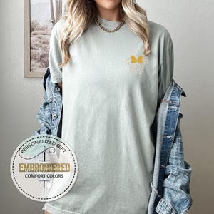 Pu&ograve; includere: Una maglietta verde chiaro con un design a farfalla gialla ricamato sul petto sinistro. La maglietta ha una vestibilit&agrave; rilassata e un collo rotondo. L'immagine include anche un logo per un'azienda chiamata "Embroidered Comfort Colors" specializzata in regali personalizzati.