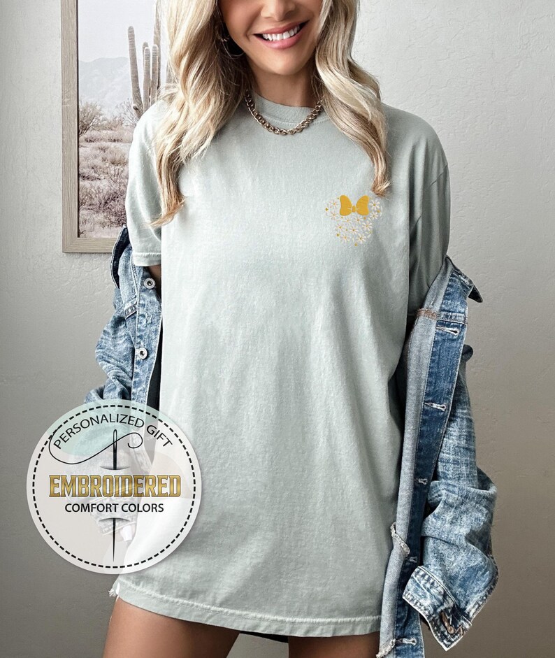 Pu&ograve; includere: Una maglietta verde chiaro con un design a farfalla gialla ricamato sul petto sinistro. La maglietta ha una vestibilit&agrave; rilassata e un collo rotondo. L'immagine include anche un logo per un'azienda chiamata "Embroidered Comfort Colors" specializzata in regali personalizzati.