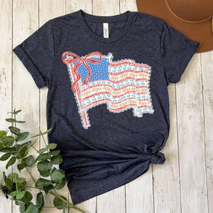 May include: A dark heather grey short-sleeve t-shirt featuring a graphic of a waving American flag. The flag design is composed of a blue field with white stars and red and white stripes, accented by a prominent red bow at the top left corner.