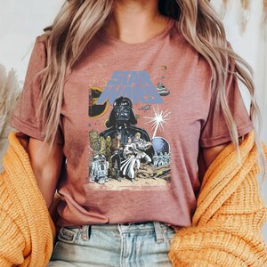 May include: Dusty rose t-shirt featuring a Star Wars graphic. The design includes Darth Vader, characters, spaceships, and the Star Wars logo in blue. The shirt is a short-sleeved crew neck.