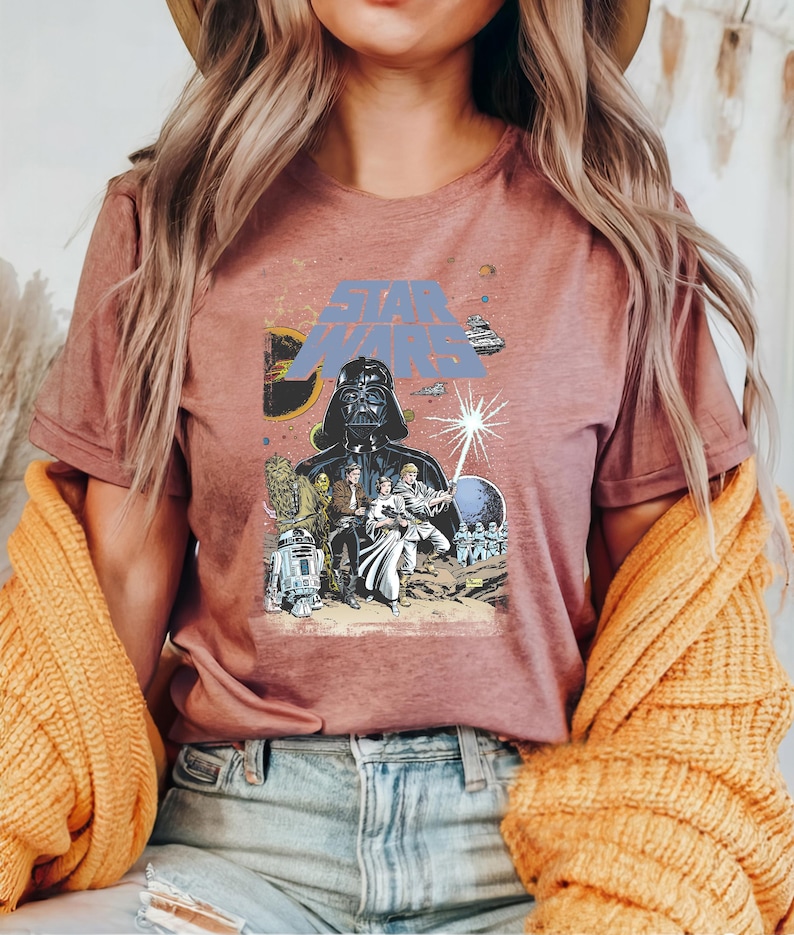 May include: Dusty rose t-shirt featuring a Star Wars graphic. The design includes Darth Vader, characters, spaceships, and the Star Wars logo in blue. The shirt is a short-sleeved crew neck.