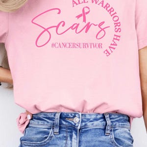 May include: Pink t-shirt with the text "All Warriors Have Scars" and a pink ribbon. The text "#cancersurvivor" is below the main text. The model is wearing blue jeans.