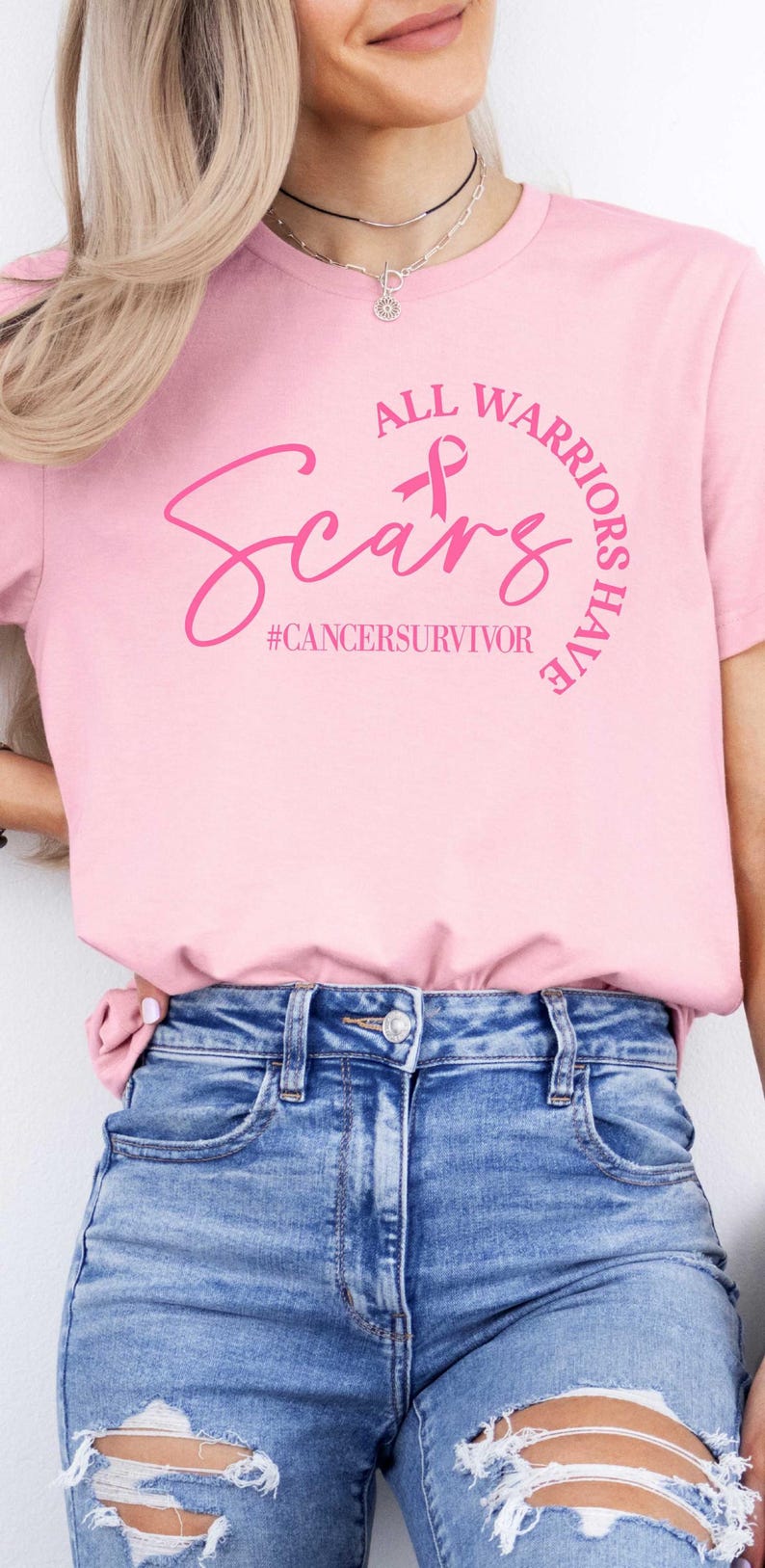 May include: Pink t-shirt with the text "All Warriors Have Scars" and a pink ribbon. The text "#cancersurvivor" is below the main text. The model is wearing blue jeans.