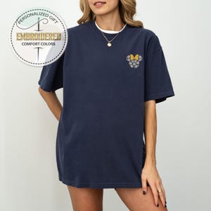 Pu&ograve; includere: Una maglietta blu navy con un ricamo floreale a forma di testa di topo con un fiocco giallo.