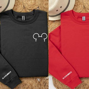 以下が含まれることがあります： 白のミッキーマウスとミニーマウスの耳のデザインが施された、黒と赤のクルーネックのトレーナー。各トレーナーの左袖にはローマ数字のテキストXLVIIIMMCXXがあります。トレーナーは織りマットの上に表示されています。