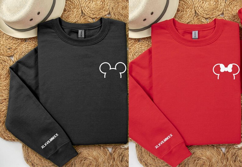 以下が含まれることがあります： 白のミッキーマウスとミニーマウスの耳のデザインが施された、黒と赤のクルーネックのトレーナー。各トレーナーの左袖にはローマ数字のテキストXLVIIIMMCXXがあります。トレーナーは織りマットの上に表示されています。