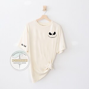 May include: Cream-coloured t-shirt featuring a black Jack Skellington face design on the front and the text "Her Jack" on the sleeve. The shirt is suspended on a wooden hanger against a white backdrop. A circular logo is visible.