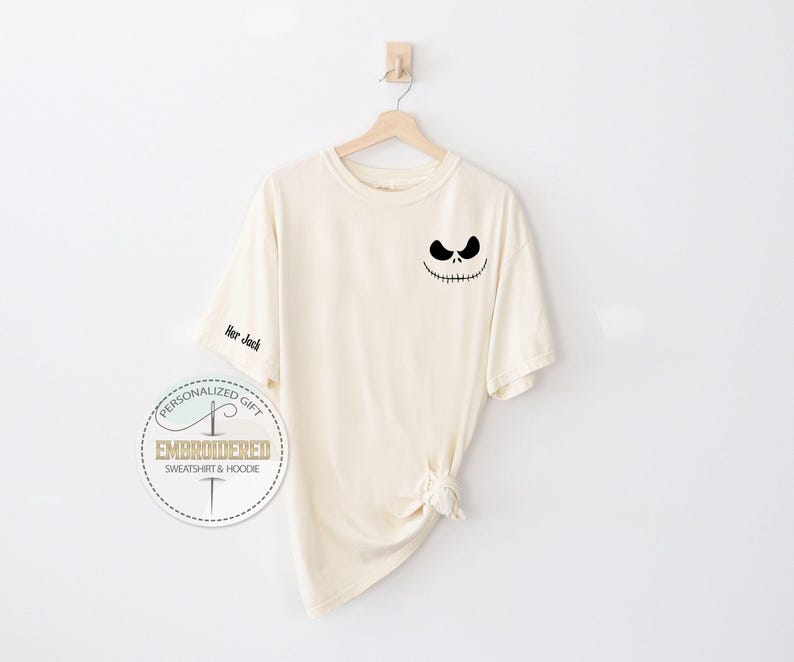 May include: Cream-coloured t-shirt featuring a black Jack Skellington face design on the front and the text "Her Jack" on the sleeve. The shirt is suspended on a wooden hanger against a white backdrop. A circular logo is visible.
