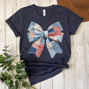 May include: A dark heather navy blue short-sleeve t-shirt featuring a large, central graphic of a decorative bow. The bow is composed of various fabric patterns including the American flag, white stars on blue, red stripes, and the Statue of Liberty.