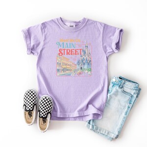 May include: A lilac-colored t-shirt with rolled sleeves featuring a Main Street graphic in pastel colors. The graphic includes a castle and buildings. The shirt is paired with denim shorts and black and white checkered slip-on shoes.