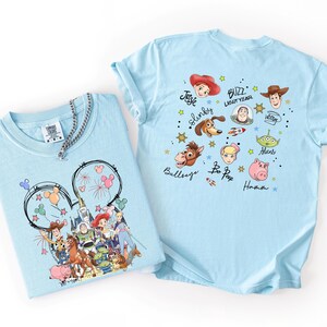 K&ouml;nnte beinhalten: Zwei hellblaue T-Shirts mit Toy Story-Charakteren. Ein Shirt hat ein Mickey Mouse-Ohren-Design mit Feuerwerk und Charakteren. Das andere Shirt zeigt Gesichter von Charakteren und Namen wie Buzz Lightyear und Woody.